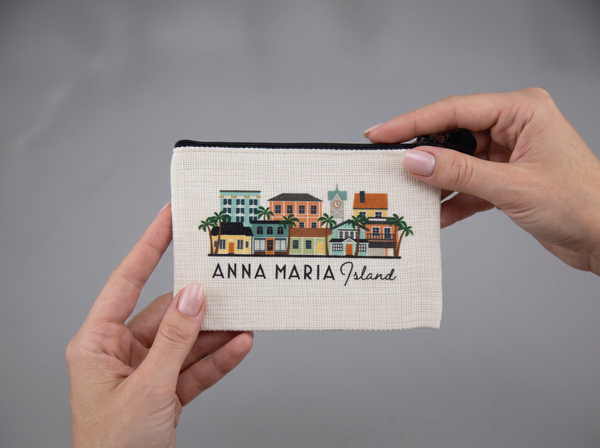 A small zippered pouch held by two hands against a neutral gray background. The pouch is off-white with a colorful embroidered illustration of buildings and palm trees, and the text 'ANNA MARIA Island' printed below.
