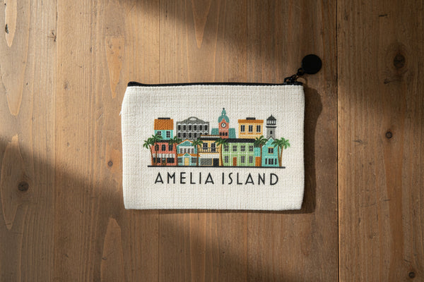A cream-colored canvas zippered pouch featuring a colorful illustration of Amelia Island buildings and the text 'AMELIA ISLAND' in black, with a black zipper and black tassel charm.