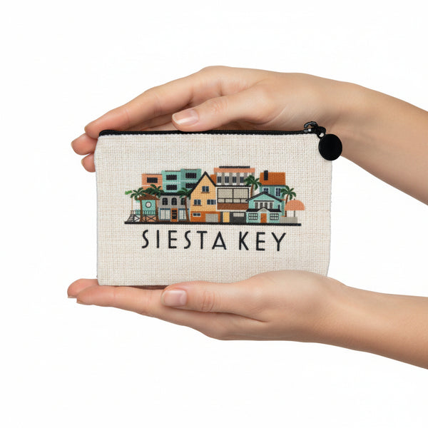An off-white canvas zippered pouch featuring a colorful embroidered illustration of buildings and palm trees from Siesta Key, with 'SIESTA KEY' embroidered in black below the artwork. A black zipper pull is attached on the right side.