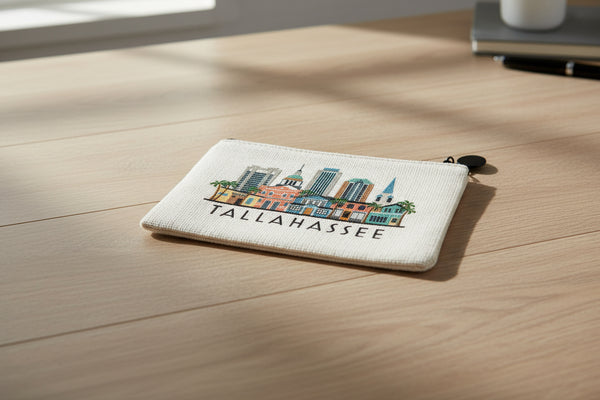 A small zip pouch with an off-white woven texture, featuring a colorful Tallahassee skyline illustration and the word 'TALLAHASSEE' printed in black. It has a black zipper and a black pom-pom charm.