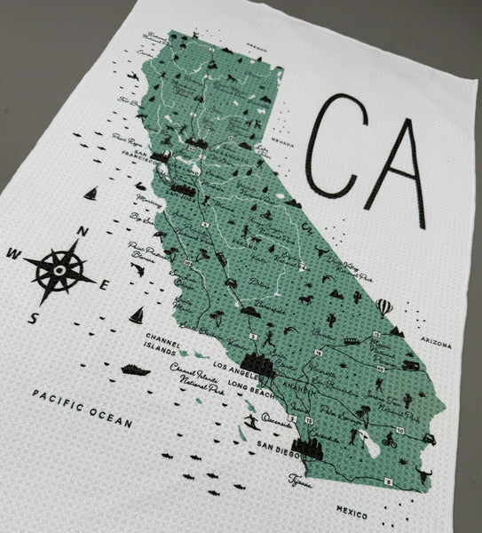 A white woven tea towel featuring a green and black map of California with the letters 'CA' printed prominently, laid flat on a neutral gray surface.