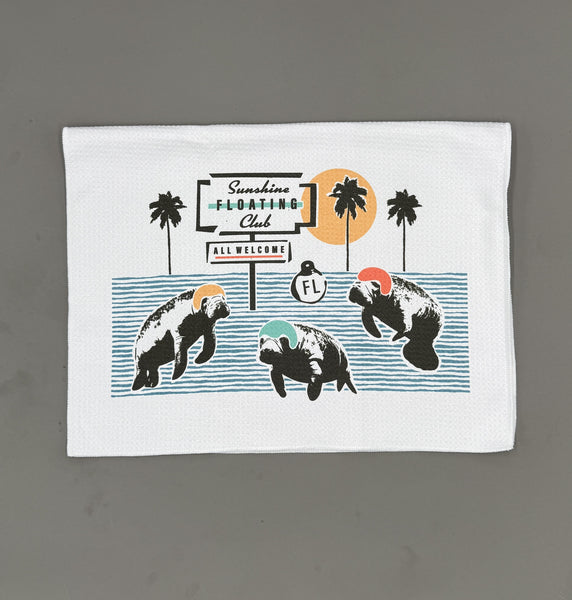 A white rectangular kitchen towel or placemat with a printed graphic of three manatees wearing colorful hats in front of palm trees and a sign reading 'Sunshine Floating Club' and 'All Welcome FL'.