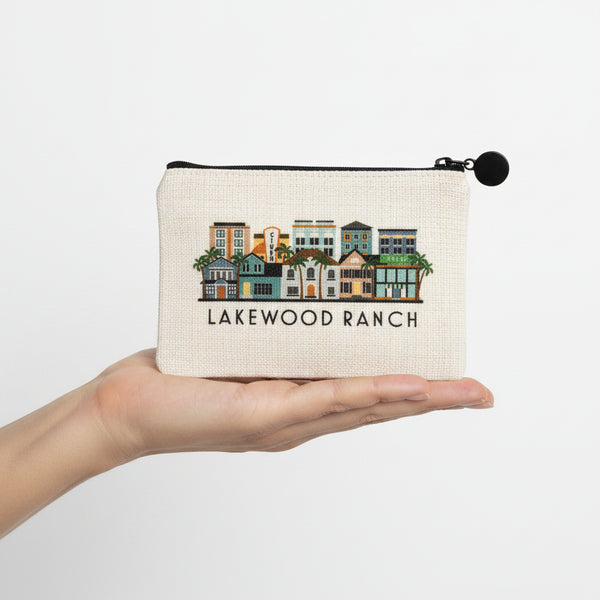 An off-white canvas zippered pouch featuring a colorful illustration of vintage houses and the text 'LAKEWOOD RANCH' below it, with a black zipper and black pom-pom pull.