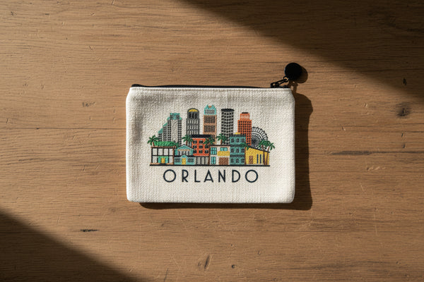 A small zip pouch with a cream-colored canvas background featuring a colorful illustrated skyline of Orlando, Florida, with the word 'ORLANDO' printed below. The pouch has a black zipper and a black pom-pom charm on the zipper pull.