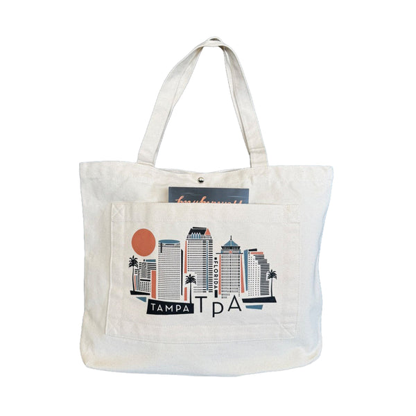 An off-white canvas tote bag featuring a Tampa skyline graphic printed on the front pocket, with a hangtag visible at the top, set against a plain white background.