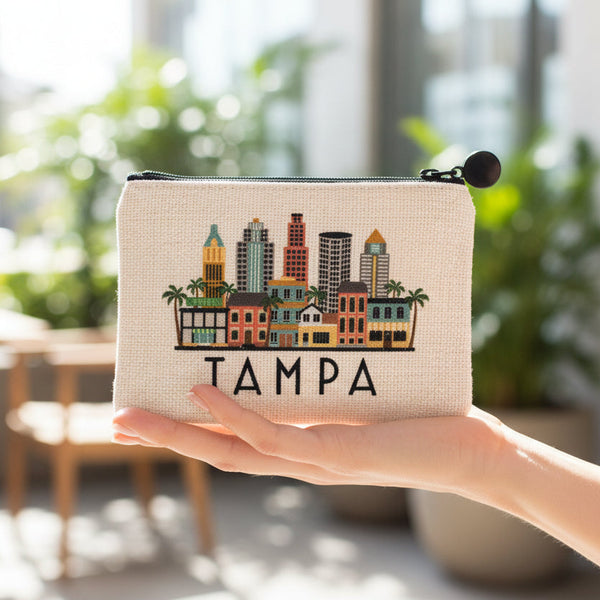 A small zip pouch with a black zipper and black pom-pom charm, featuring a colorful Tampa skyline illustration and the word 'TAMPA' printed below, on a white background.