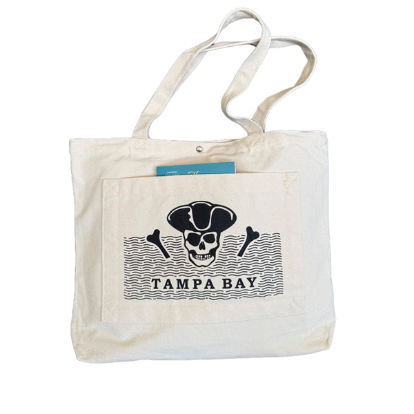 An off-white canvas tote bag with a front pocket featuring a black pirate skull and crossbones graphic above the text 'TAMPA BAY'.