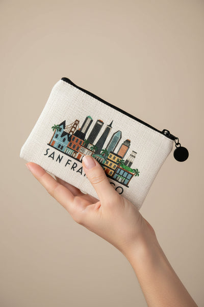 A small canvas pouch with a black zipper and black pom-pom pull, featuring a colorful San Francisco skyline illustration and the text 'SAN FRANCISCO' on a light beige background.