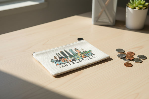 A small canvas pouch with a black zipper and black pom-pom pull, featuring a colorful San Francisco skyline illustration and the text 'SAN FRANCISCO' on a light beige background.